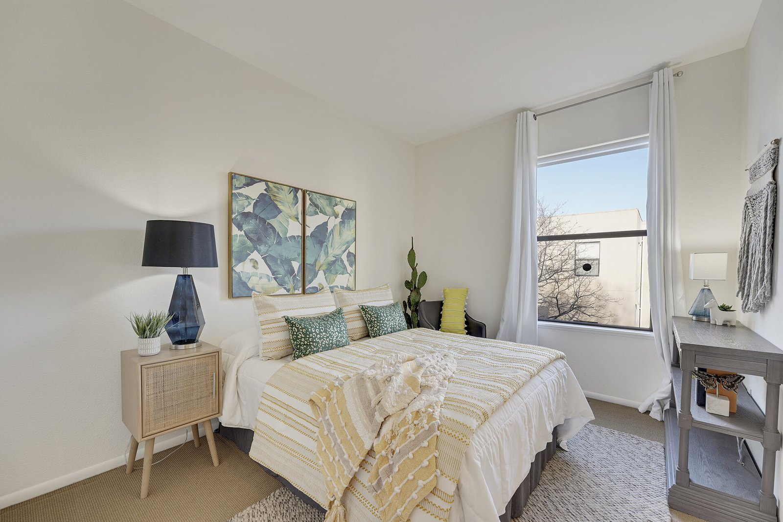 Modern bedroom with a cactus in the corner, a bed with green and yellow accents, and a large window showing a neighboring building.