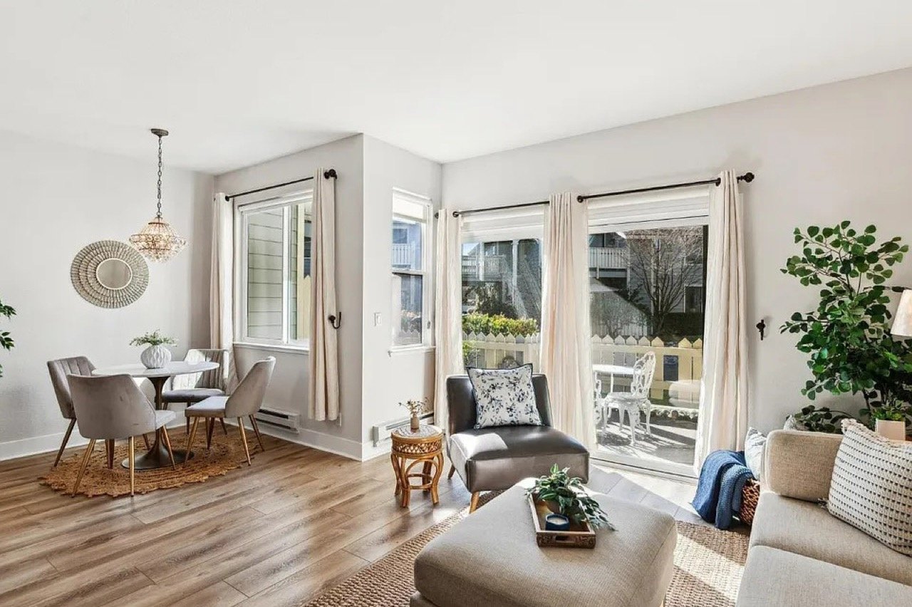Modern bright living room and dining area with light wood floors, a round white table with grey upholstered chairs, and large windows with white curtains.